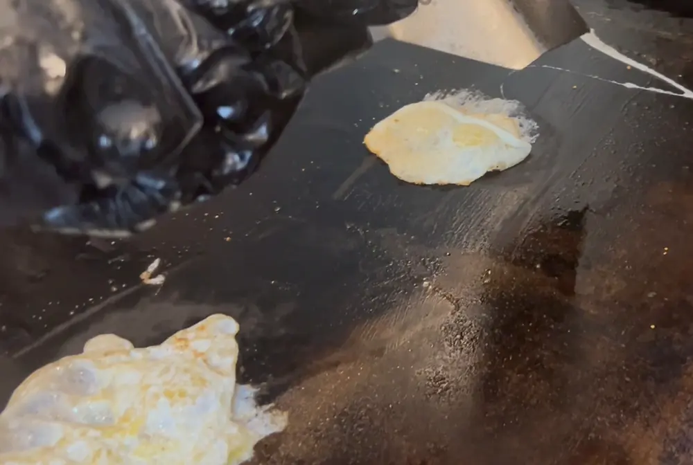A gloved hand turning small fried quail eggs on a well seasoned flat top griddle with a metal spatula.